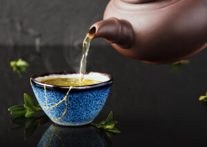 Hot green tea is poured from a ceramic teapot into a cup that has cracked and been repaired. 